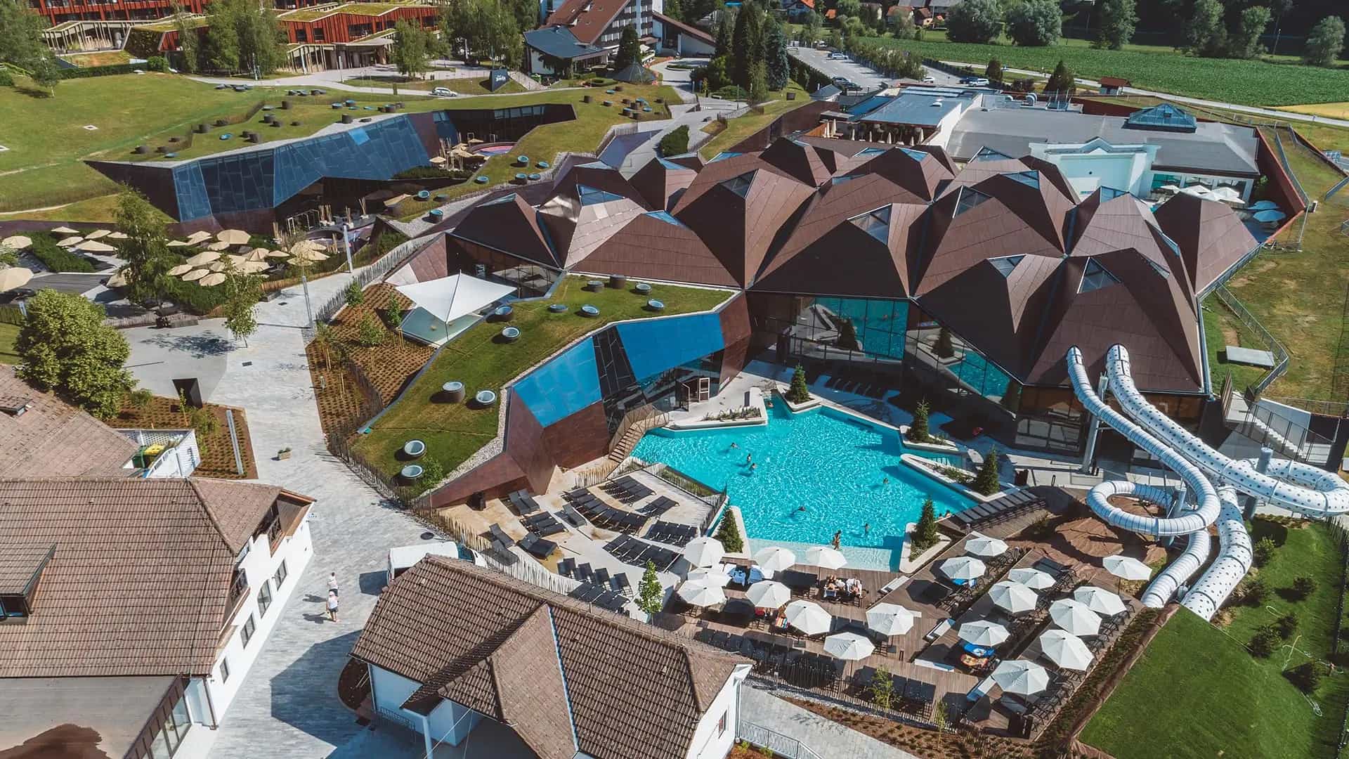 Aerial view of modern spa complex with geometric brown roof, blue pool, and white water slides.