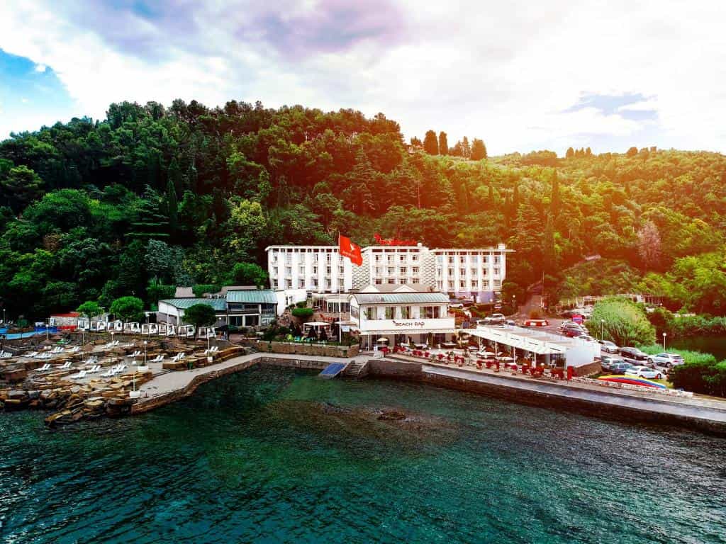 Hotel complex with beach bar nestled against a dense green hillside above turquoise coastal water.