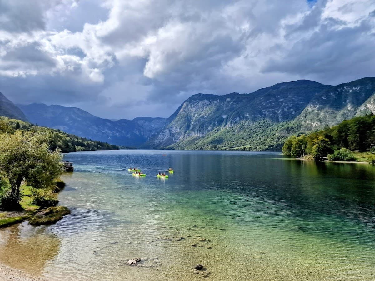 Kayakers paddling on clear alpine lake surrounded by steep, forested mountains under cloudy sky