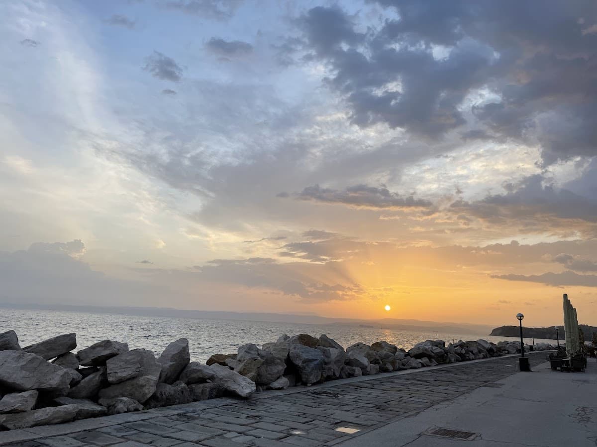 Sunset over the sea from a stone-lined promenade with a breakwater of large rocks.