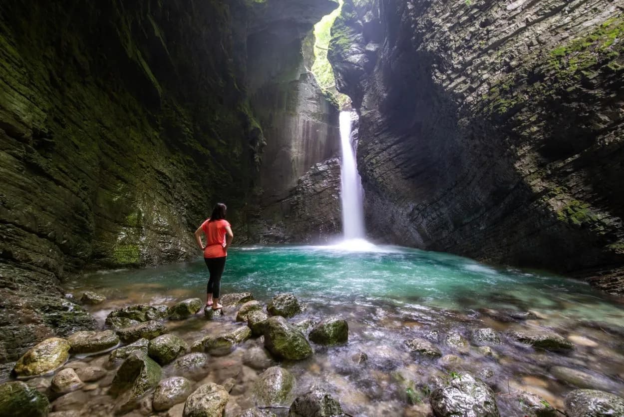 Woman observing Kozjak Waterfall plunging into turquoise pool in mossy canyon.