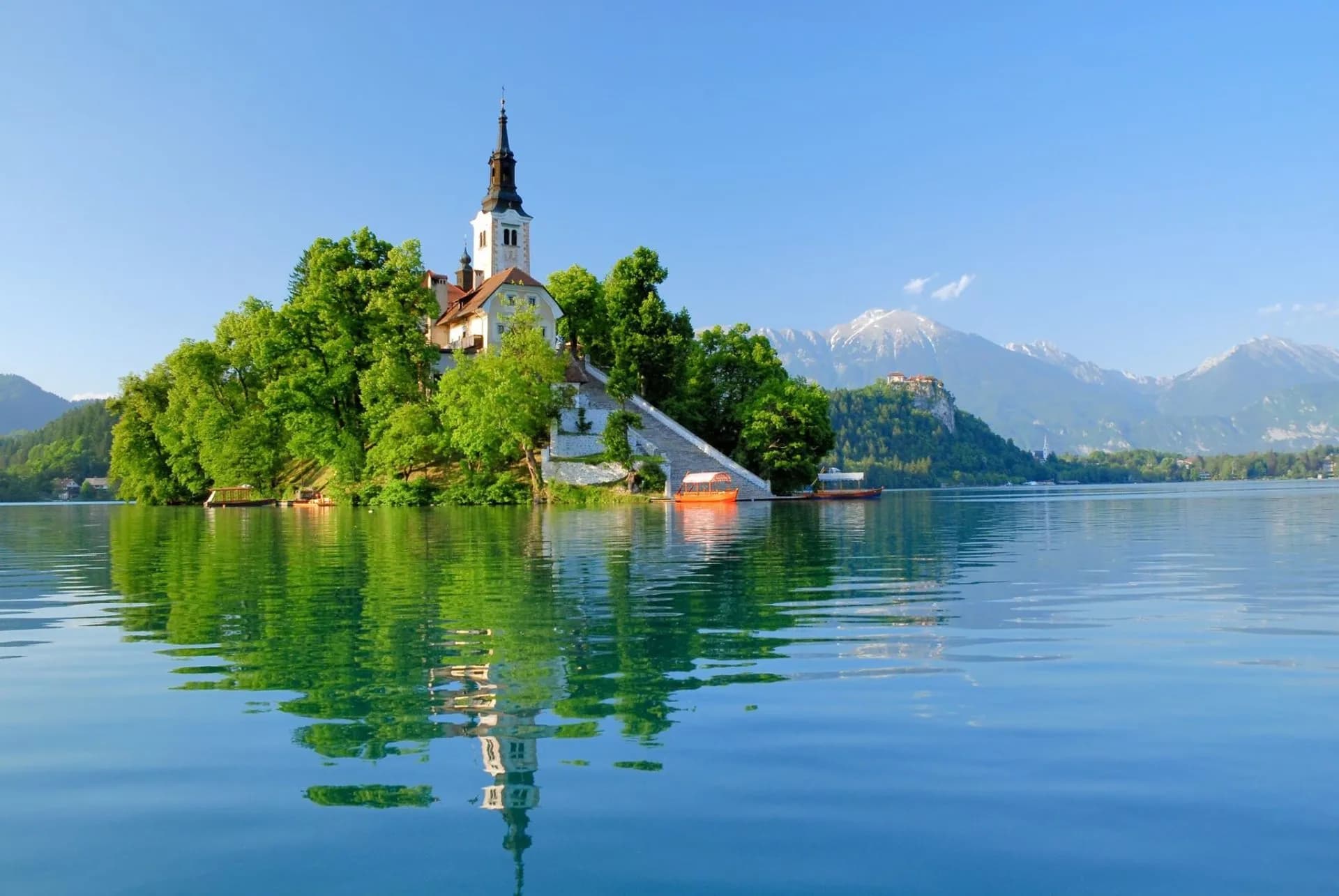 Bled Island church reflected on Lake Bled with boats and snow-capped mountains.