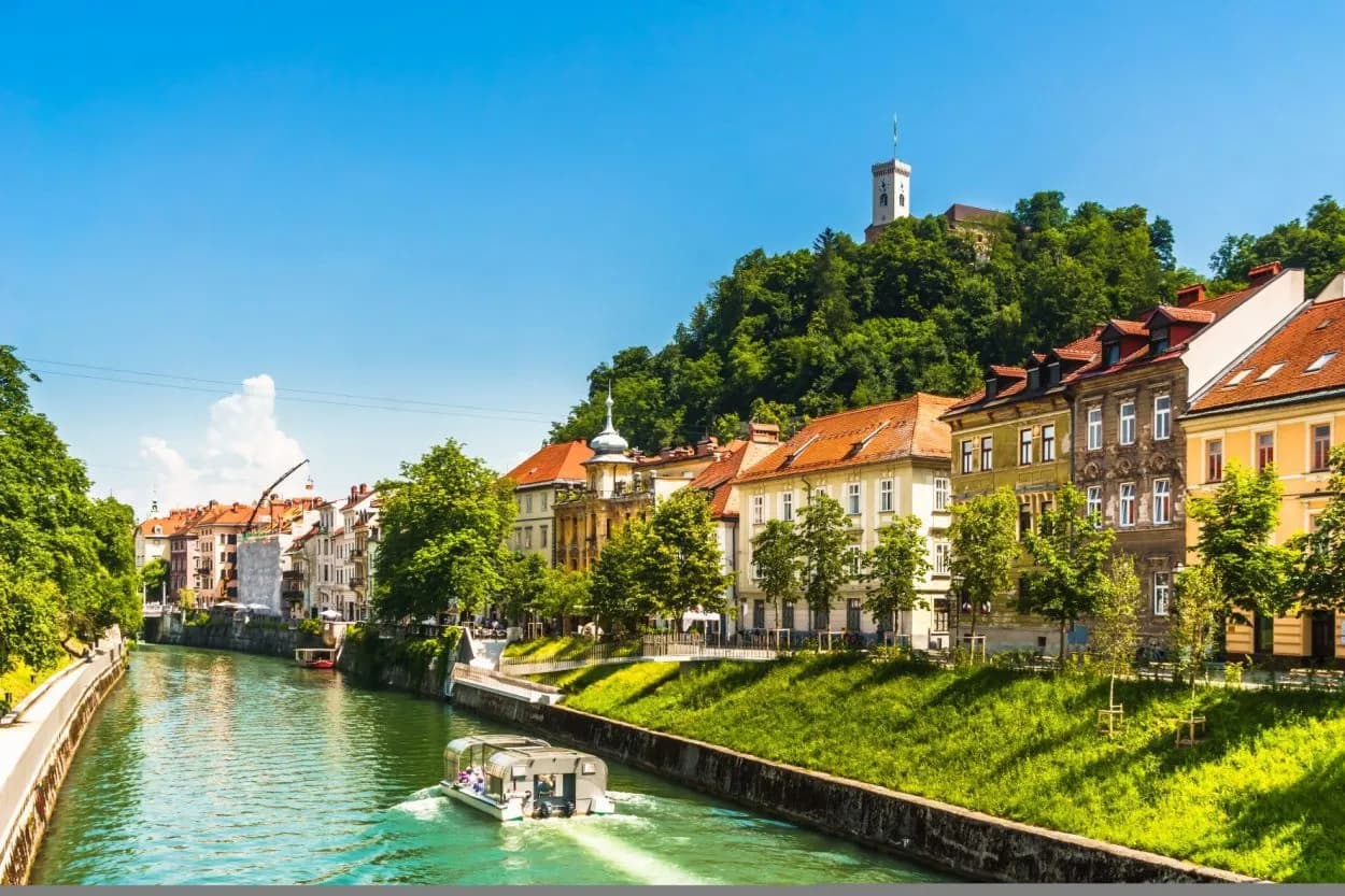 Tour boat on Ljubljanica River past colorful buildings toward Ljubljana Castle hill.