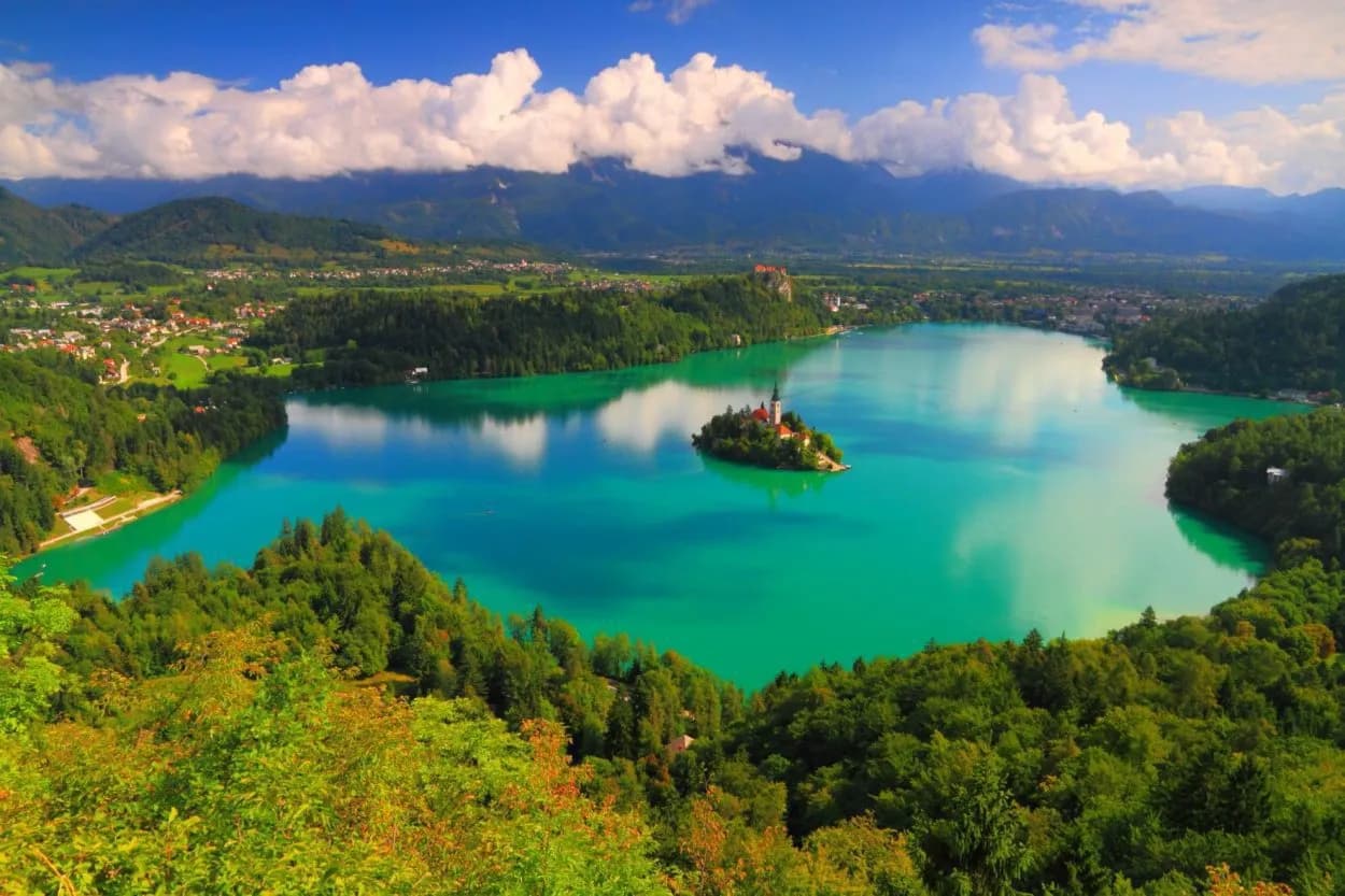 Lake Bled island church surrounded by turquoise water, green hills, and mountains under a blue sky.