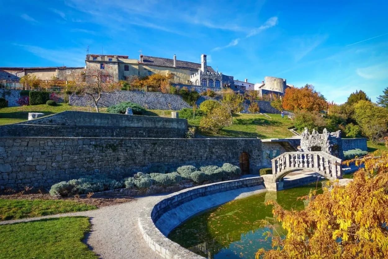 Terraced gardens with stone walls, arched bridge, and buildings atop a hill under a blue sky.