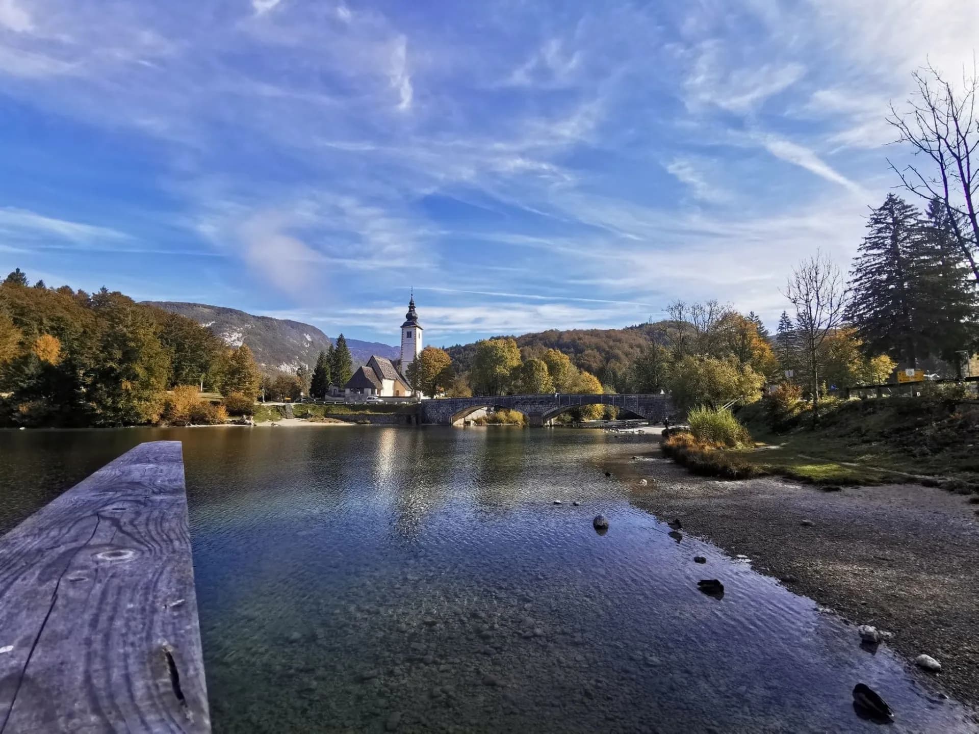 Church next to Lake Bohinj with stone bridge, autumn trees, and clear water.