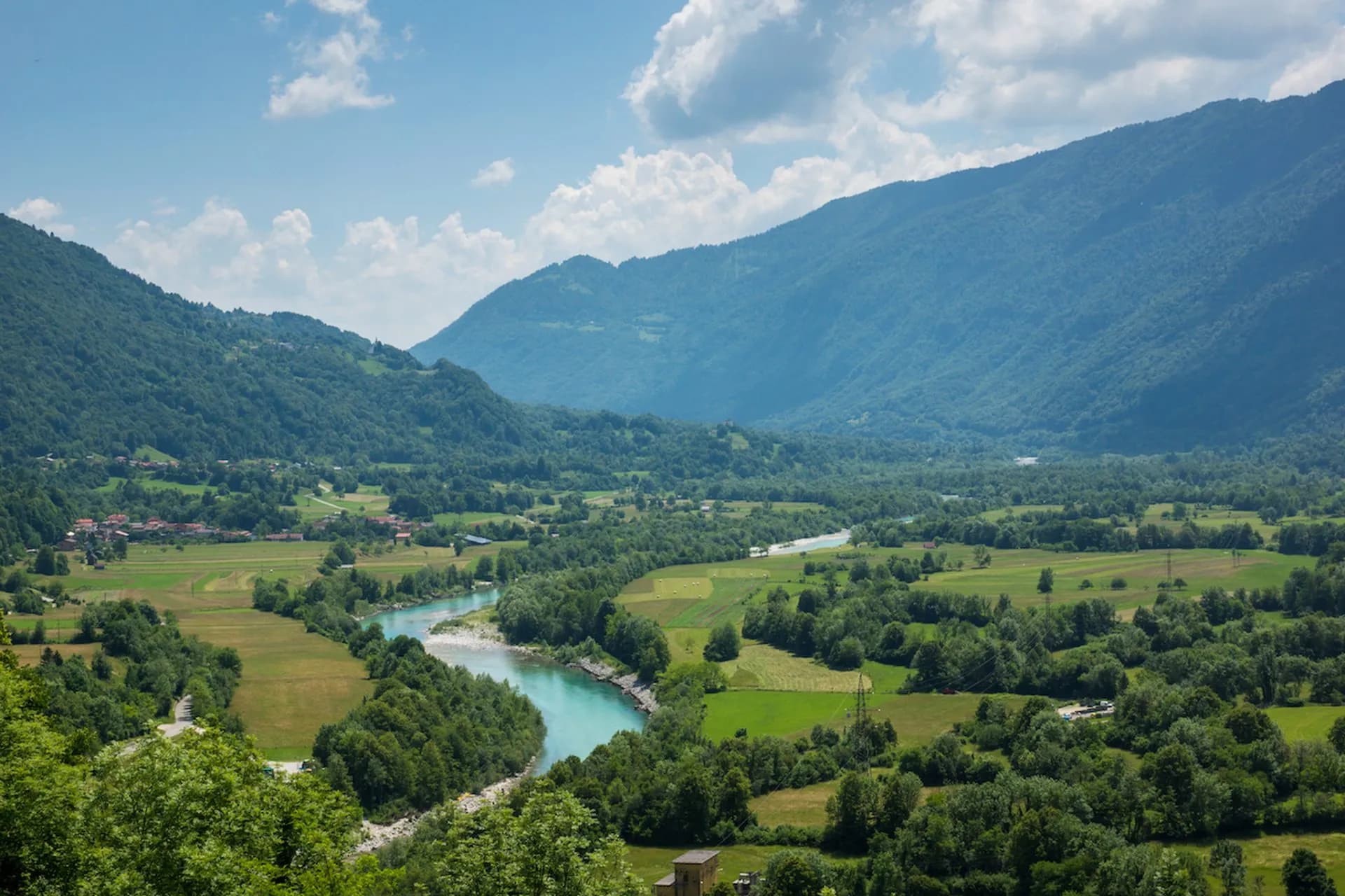 River valley with turquoise water winding through green fields and forested mountains under a blue sky.