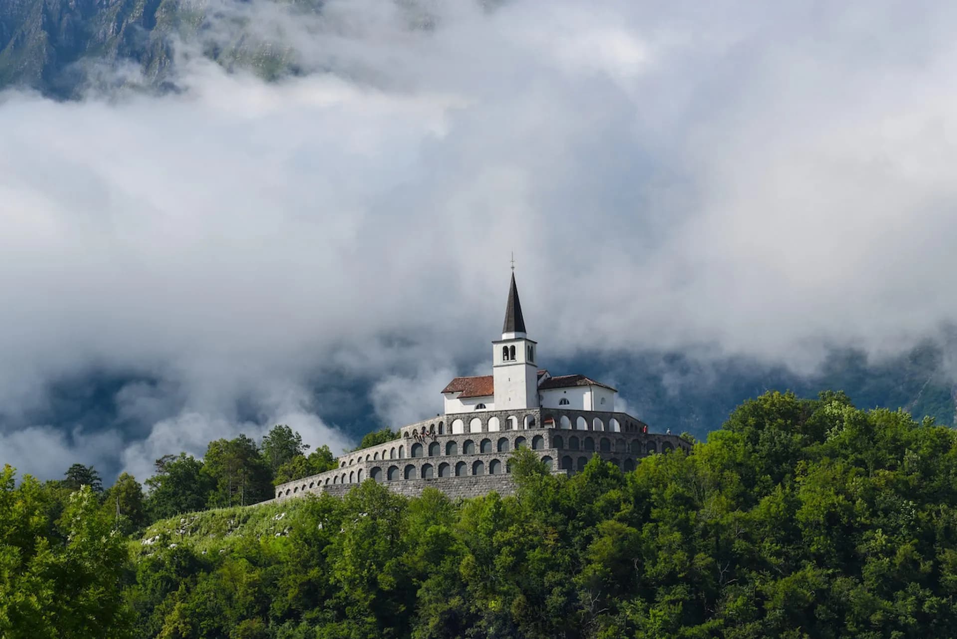 WWI memorial church on a terraced hill surrounded by lush green trees and low clouds in Kobarid.