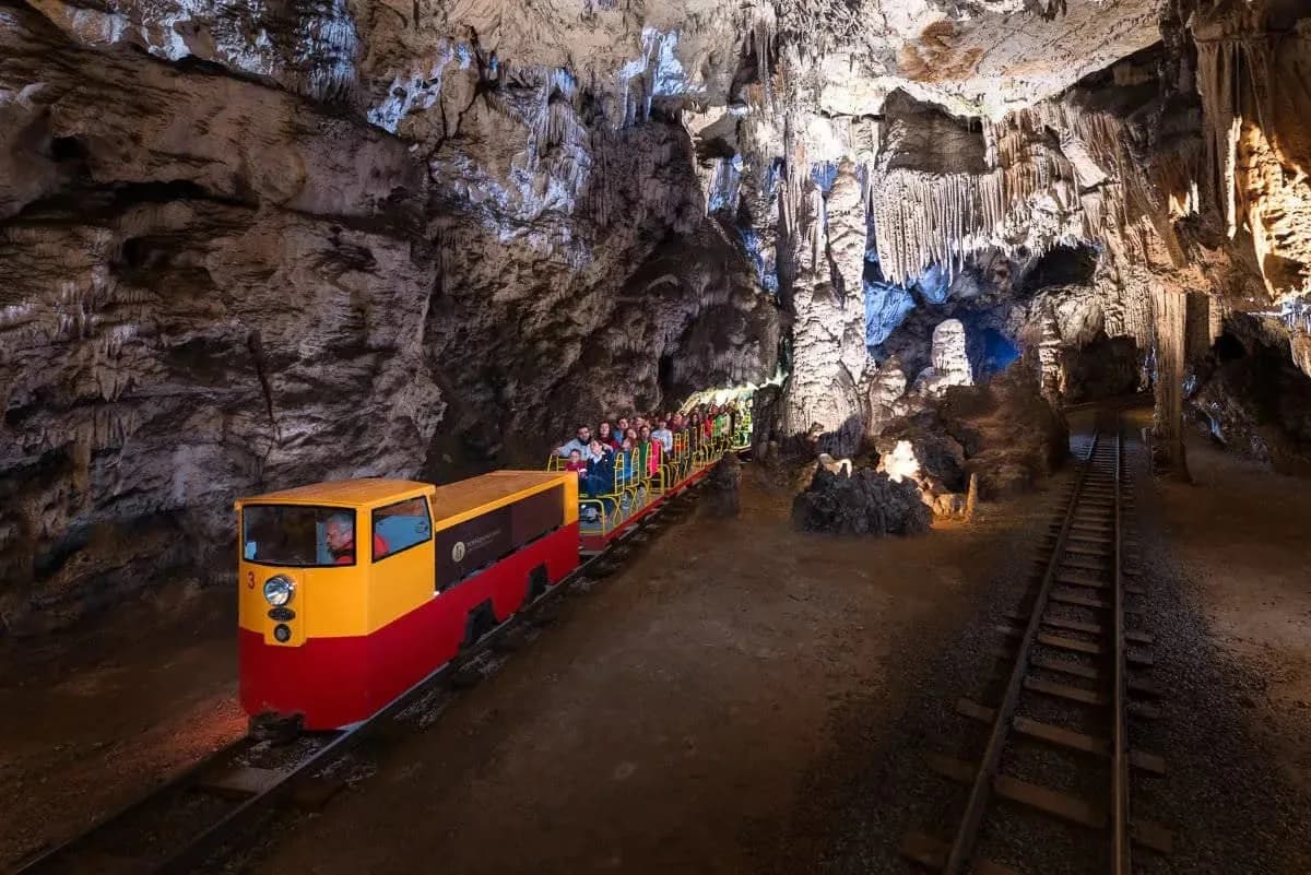 Train carrying tourists through Postojna Cave past illuminated stalactites and rock formations.