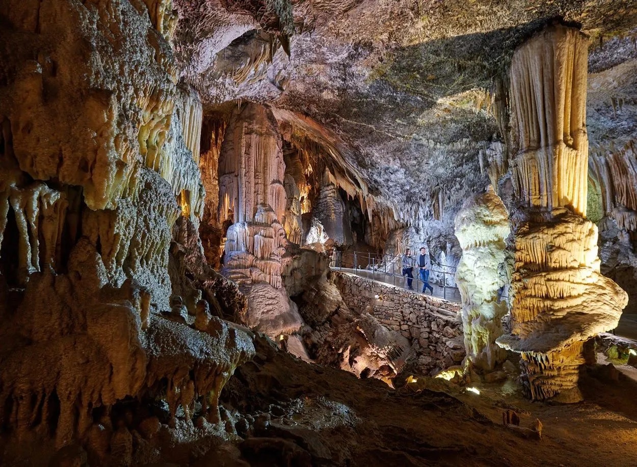 Tourists walking along a path inside Postojna Cave among massive illuminated stalactites and stalagmites.