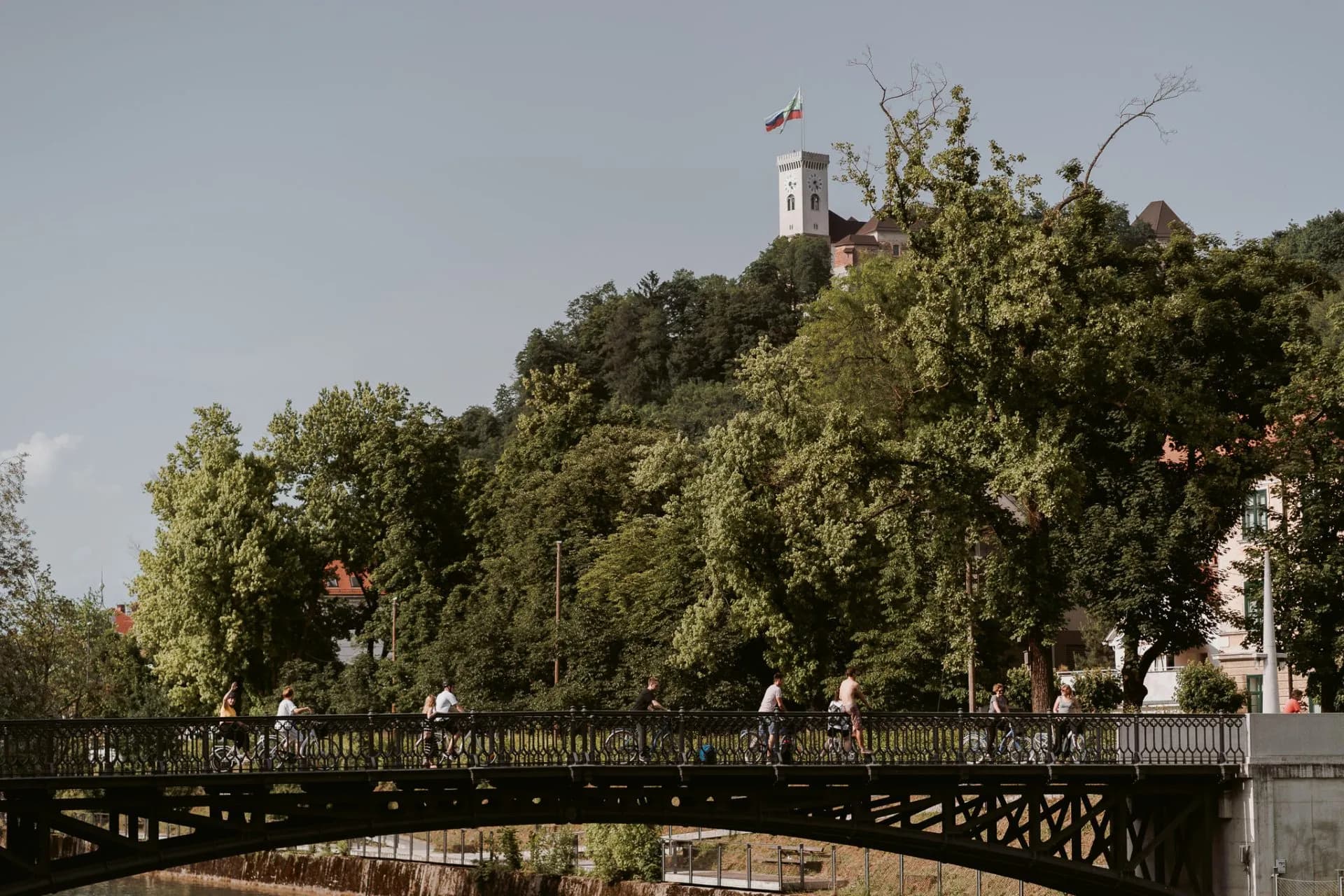Cyclists crossing a bridge with Ljubljana Castle tower visible over green trees.