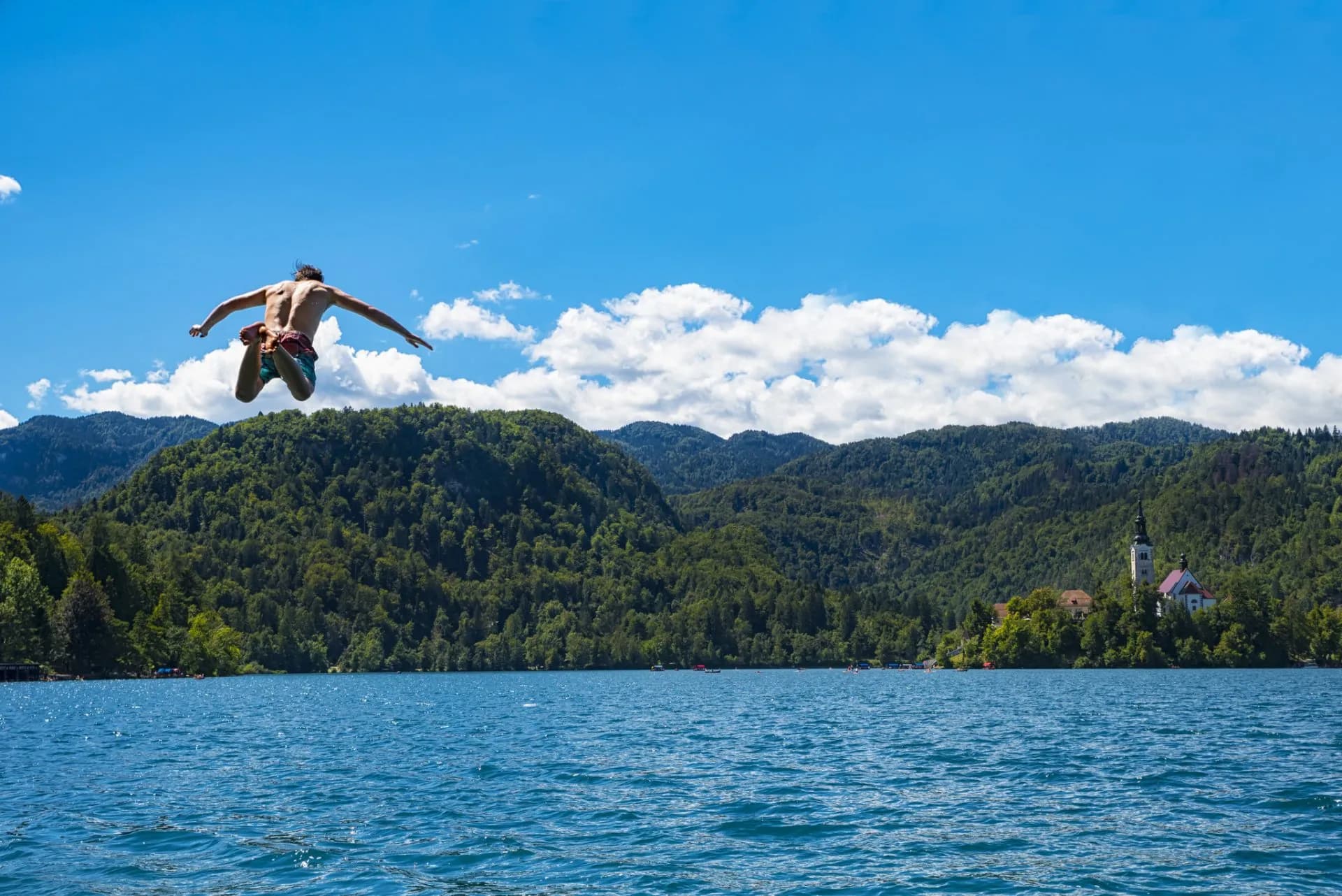 Man jumping into Lake Bled with church on island and forested mountains background