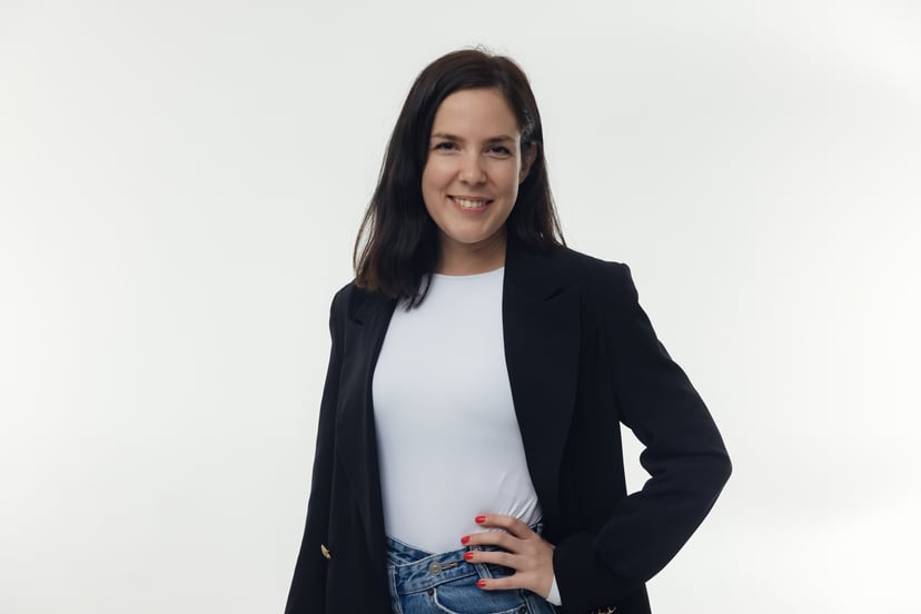 Woman with dark hair smiling while wearing a white t-shirt, blue jeans, and a black blazer against a white background.
