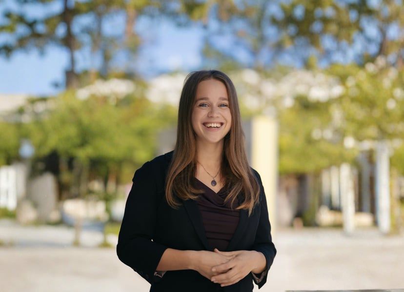 Smiling woman in a black blazer standing outdoors with a blurred park background.